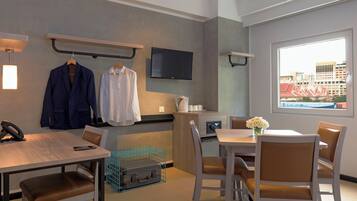 A hotel room with a dining area, a TV mounted on the wall, a window with a city view, and a desk with a phone and a suitcase.