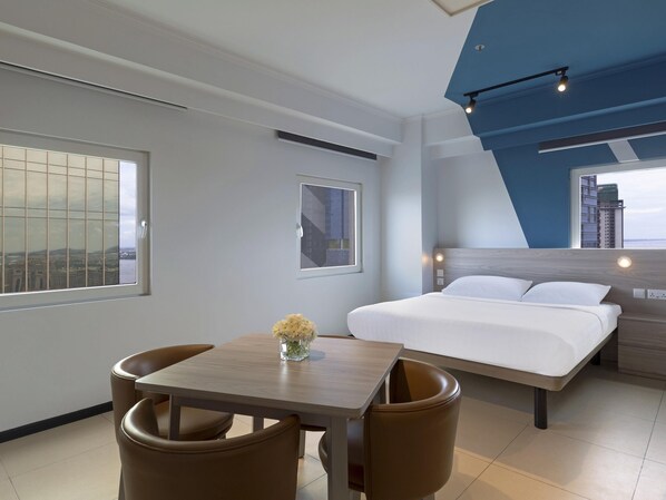 Habitación de hotel moderna con cama, una mesa de comedor con sillas y vistas a la ciudad a través de amplias ventanas.