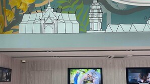 Un mostrador de recepción con dos monitores, una persona trabajando en un ordenador portátil y un mural con un templo y un elefante.