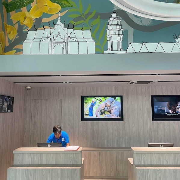 Un comptoir d’accueil avec deux écrans d’ordinateur, une personne travaillant sur un ordinateur portable, et une fresque représentant un temple et un éléphant.