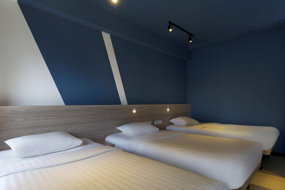 Habitació d'hotel amb tres llits, un panell de capçalera de fusta i un lluminari modern.