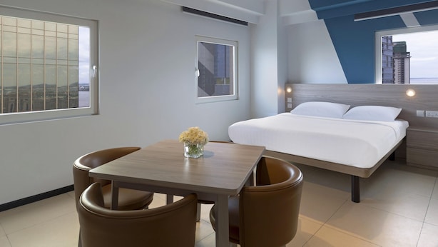 Habitación de hotel moderna con cama, una mesa de comedor con sillas y vistas a la ciudad a través de amplias ventanas.