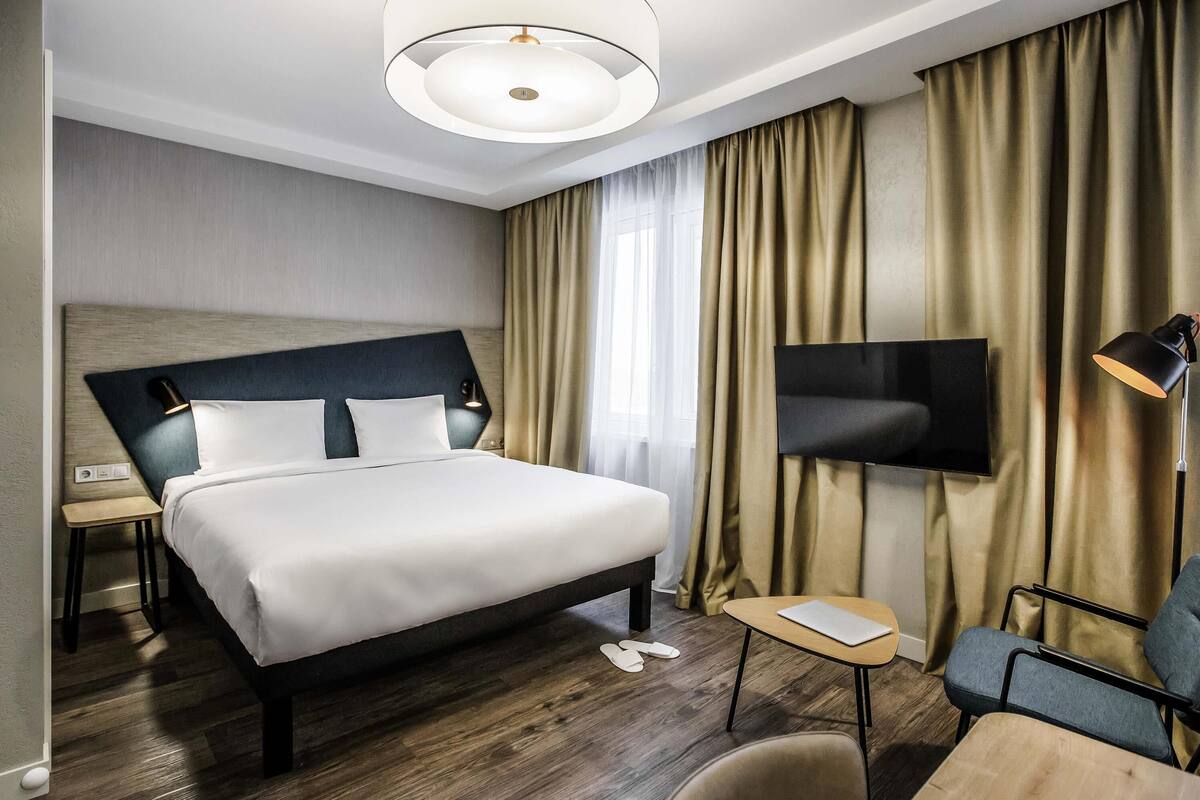 A hotel room with a large bed, a desk, a chair, a TV, and a lamp.