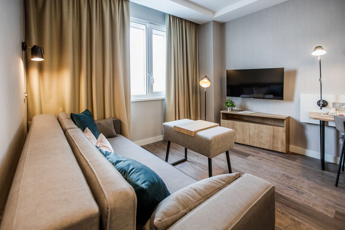 A modern living room with a sofa, ottoman, and a small table with a lamp.