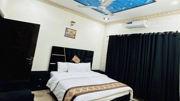 Ein Hotelzimmer mit Bett, Deckenventilator, schwarzem Sessel, schwarzem Vorhang und einer Wand-Klimaanlage.