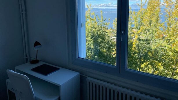 Una habitación con escritorio, silla y ventana con vistas a árboles y un cuerpo de agua.
