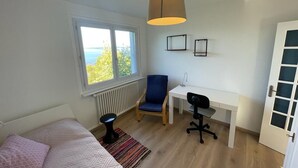 Un dormitorio con cama, escritorio, silla, radiador y ventana con vistas.