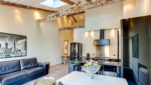 A modern living room with a sofa, a dining table with chairs, a kitchen area with a stove, and a large framed cityscape picture on the wall.