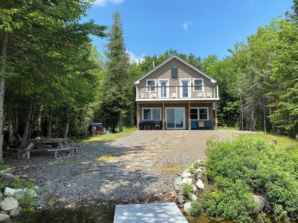 A two-story house with a balcony, surrounded by trees and a gravel path leading to the entrance.