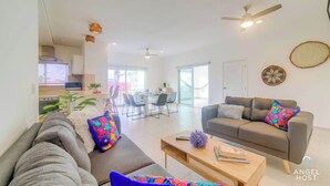 A modern living room with a grey sofa, a wooden coffee table, and a brown sofa with colorful cushions. There is a dining area in the background with a table and chairs. - Walk to 5th Ave & Beach · Pools, Balcony & Parking (Playa del Carmen)