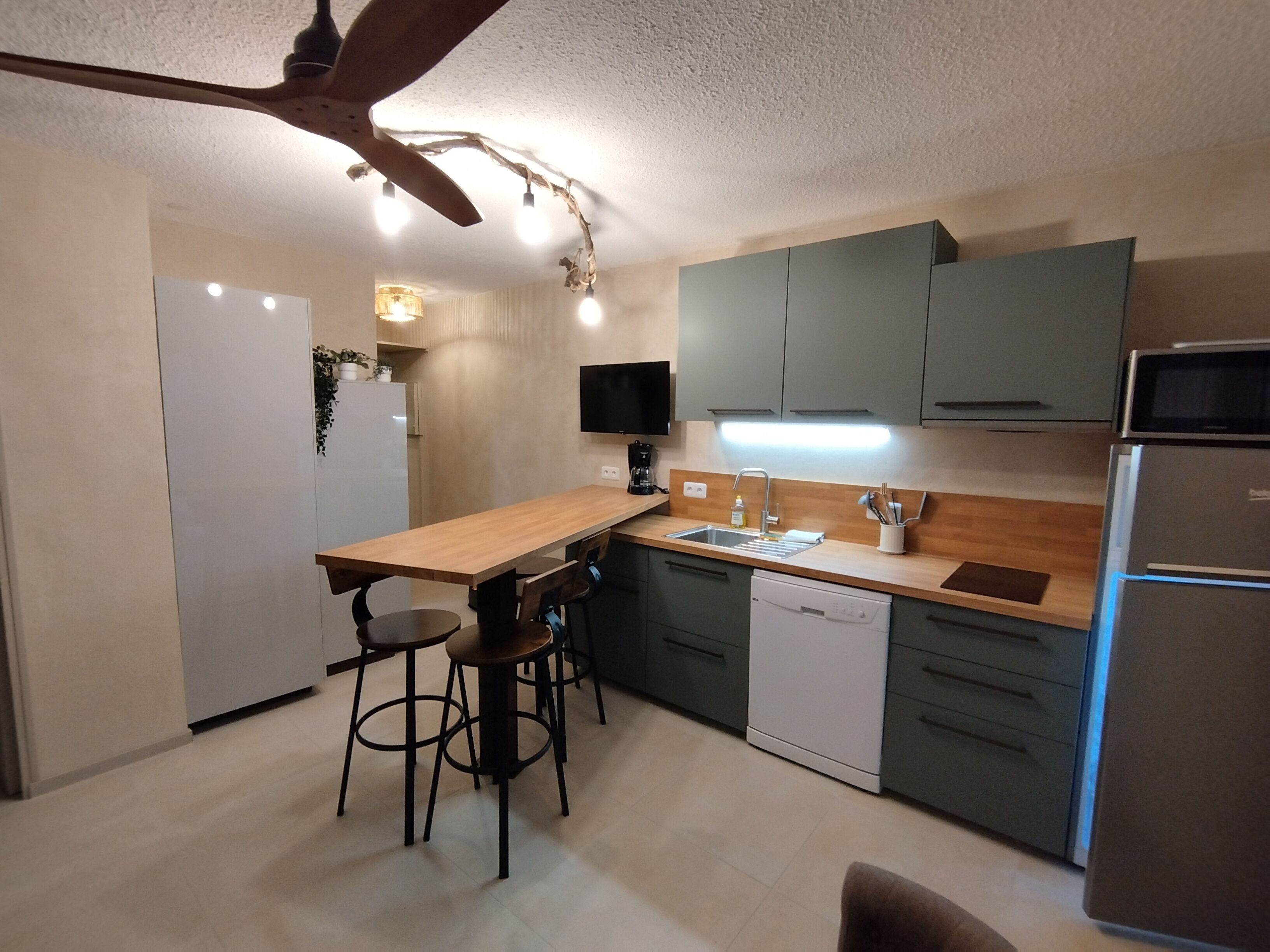 A modern kitchen with a wooden island, bar stools, a built-in dishwasher, and a flat-screen TV mounted on the wall.