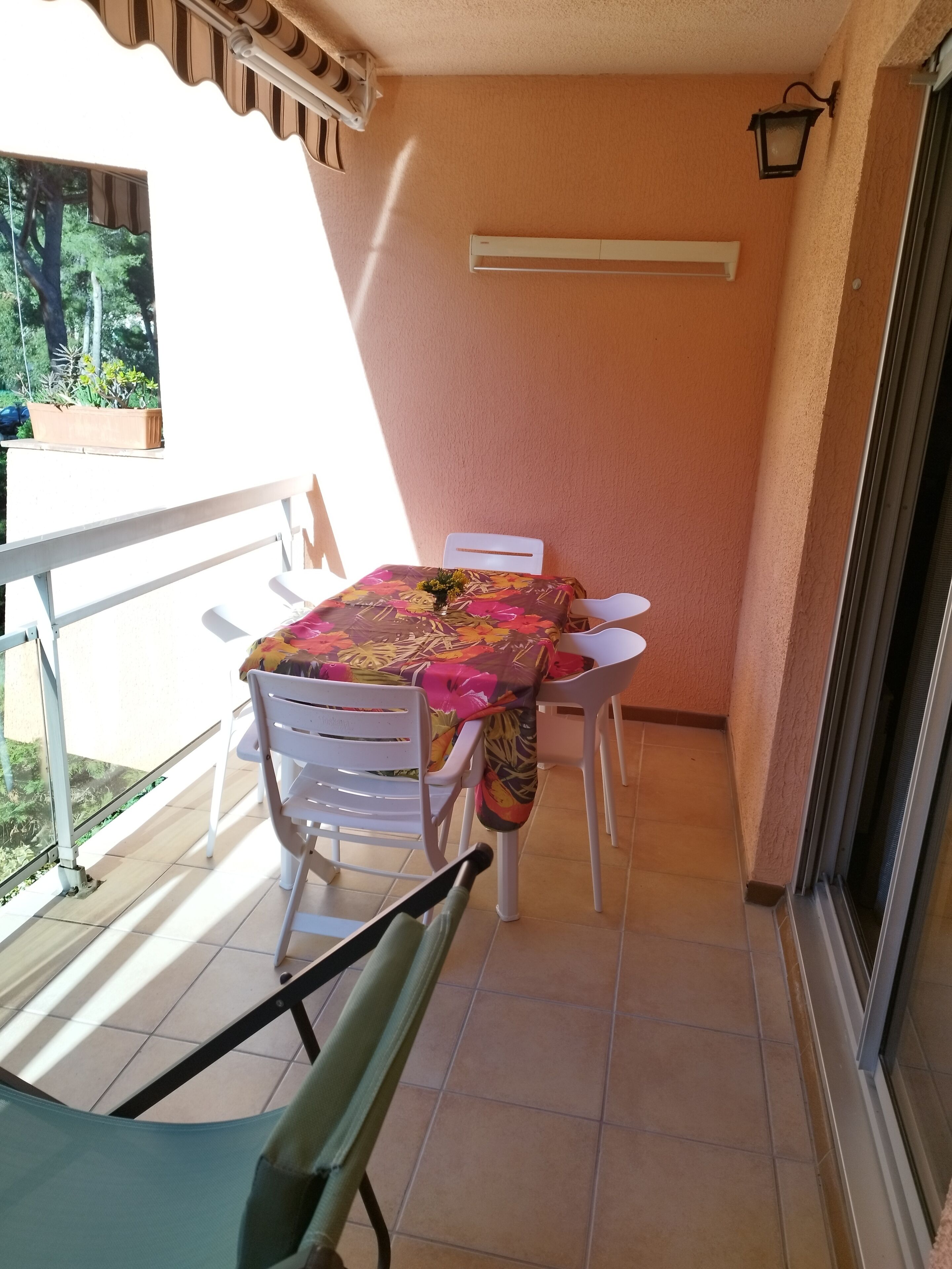 Un balcon avec une table recouverte d’une nappe à motifs floraux, des chaises blanches et une plante en pot.