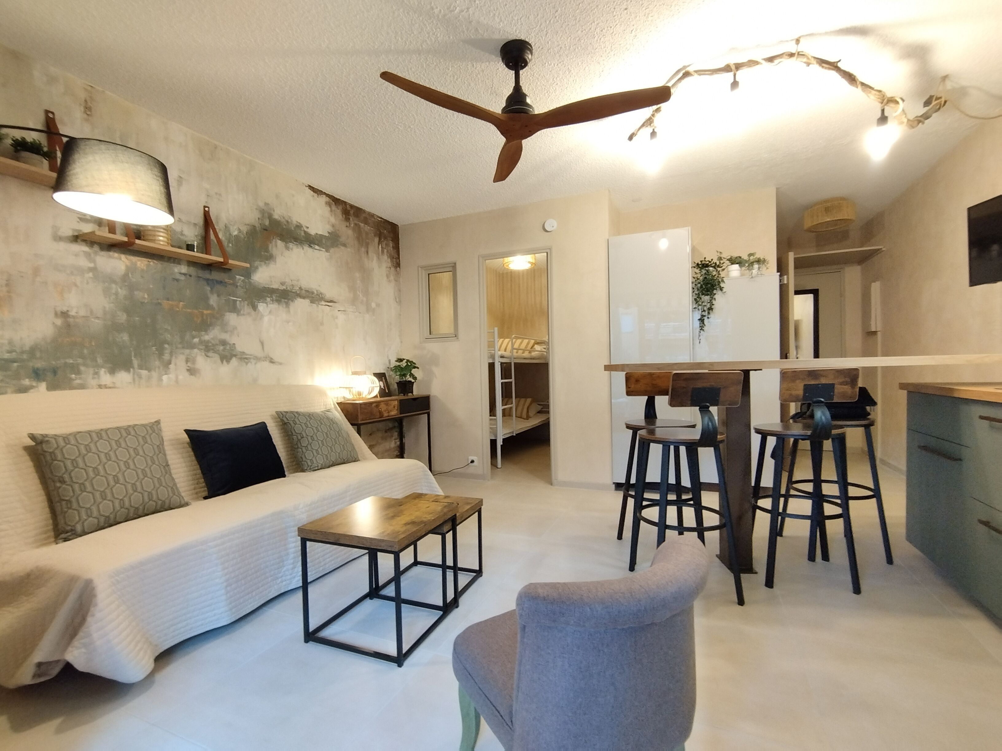 A modern living room with a sofa, coffee table, and bar stools. There is a ceiling fan, a kitchen area, and a small dining table.