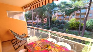 A balcony with a table covered in a floral tablecloth, white chairs, and a hammock. The balcony overlooks a garden with trees and buildings in the background. - Air-conditioned apartment 3 minutes' walk from the beach and shops (Bormes-les-Mimosas)