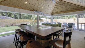 A covered outdoor dining area with a wooden table and chairs, large windows, and a view of a backyard.