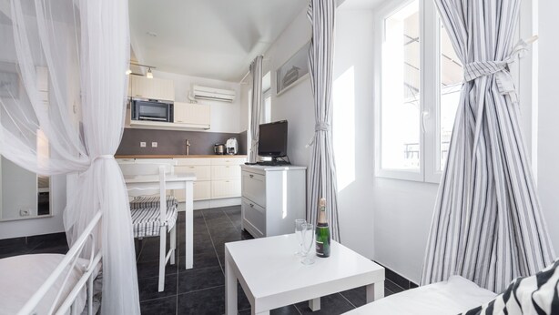A modern hotel room with a bed, a small table with a bottle and glasses, a TV, a kitchen area, and a balcony with curtains. - Florella Residences Studio terrasse Maréchal Joffre (Cannes)
