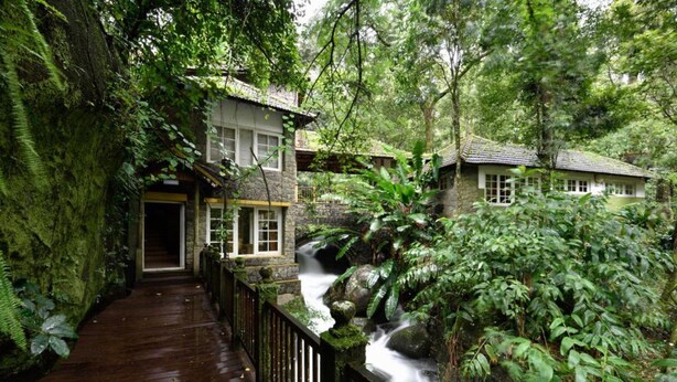A wooden pathway leading to a building surrounded by lush greenery and a waterfall. - Mystic Mayapott Thekkady (Idukki)
