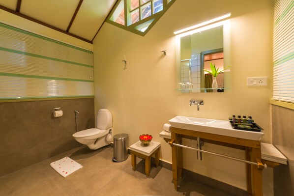 A bathroom with a toilet, sink, and mirror. There is a small table with a bowl of flowers and a trash can.
