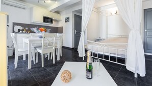 Una habitación de hotel moderna con una cama blanca, una zona de comedor con mobiliario blanco y una cocina, además de una cortina blanca sobre la cama.