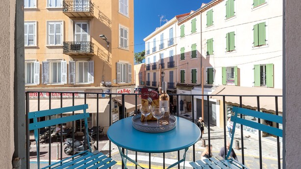 Un balcón con una mesa dispuesta para dos personas, con una botella y copas, con vistas a una calle con edificios y una tienda.