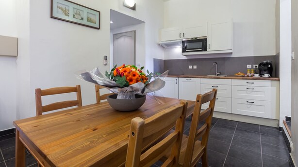 Un comedor con una mesa y sillas de madera, un centro de mesa con flores, y una cocina con armarios blancos y un microondas.