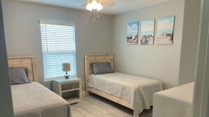 A bedroom with two single beds, a nightstand with a lamp, a ceiling fan, and a window with blinds. There are three wall-mounted pictures depicting beach scenes. - Hackberry Hideaway III (Hackberry)