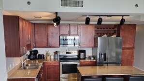 A modern kitchen with wooden cabinets, stainless steel appliances, a central island with bar stools, and a seating area with patterned cushions.