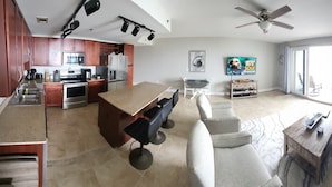 A modern kitchen with stainless steel appliances, a dining area with a wooden table and bar stools, and a living room with a sofa and a ceiling fan.