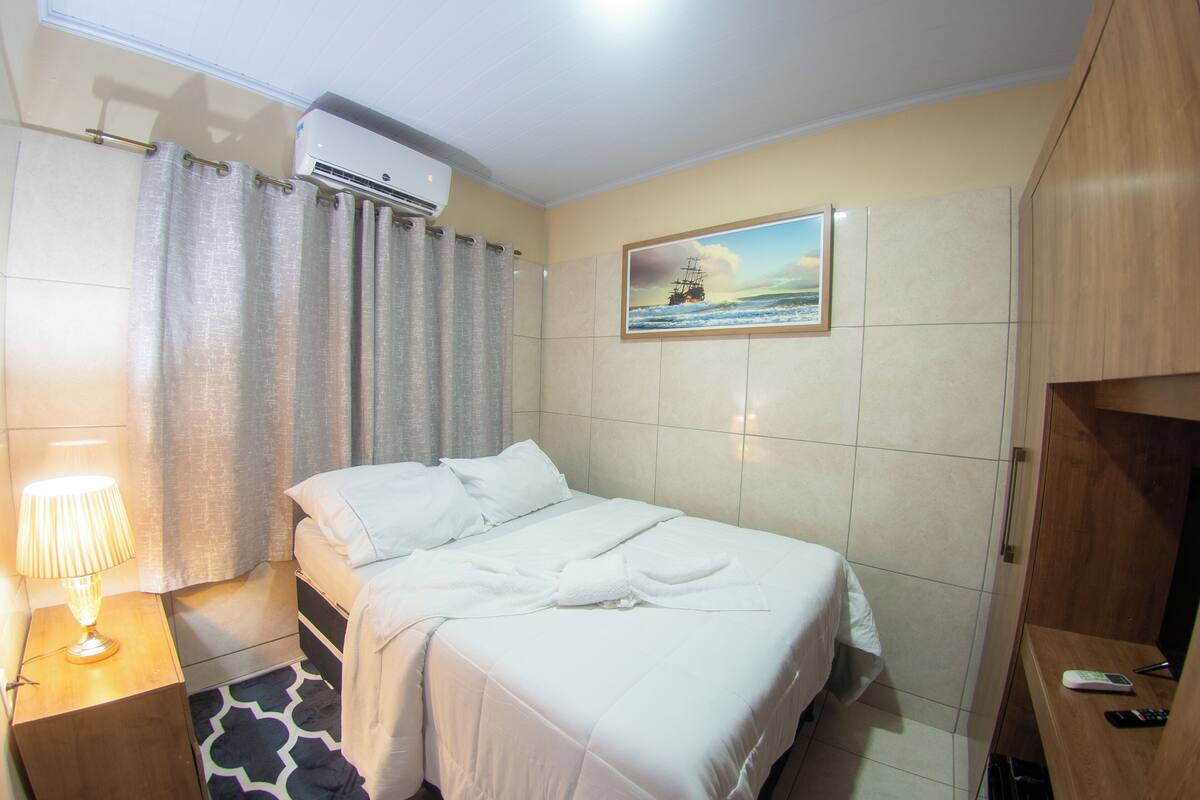 Quarto de hotel com uma cama, uma mesa de cabeceira com luminária, um guarda-roupa de madeira e uma pintura de um navio na parede.