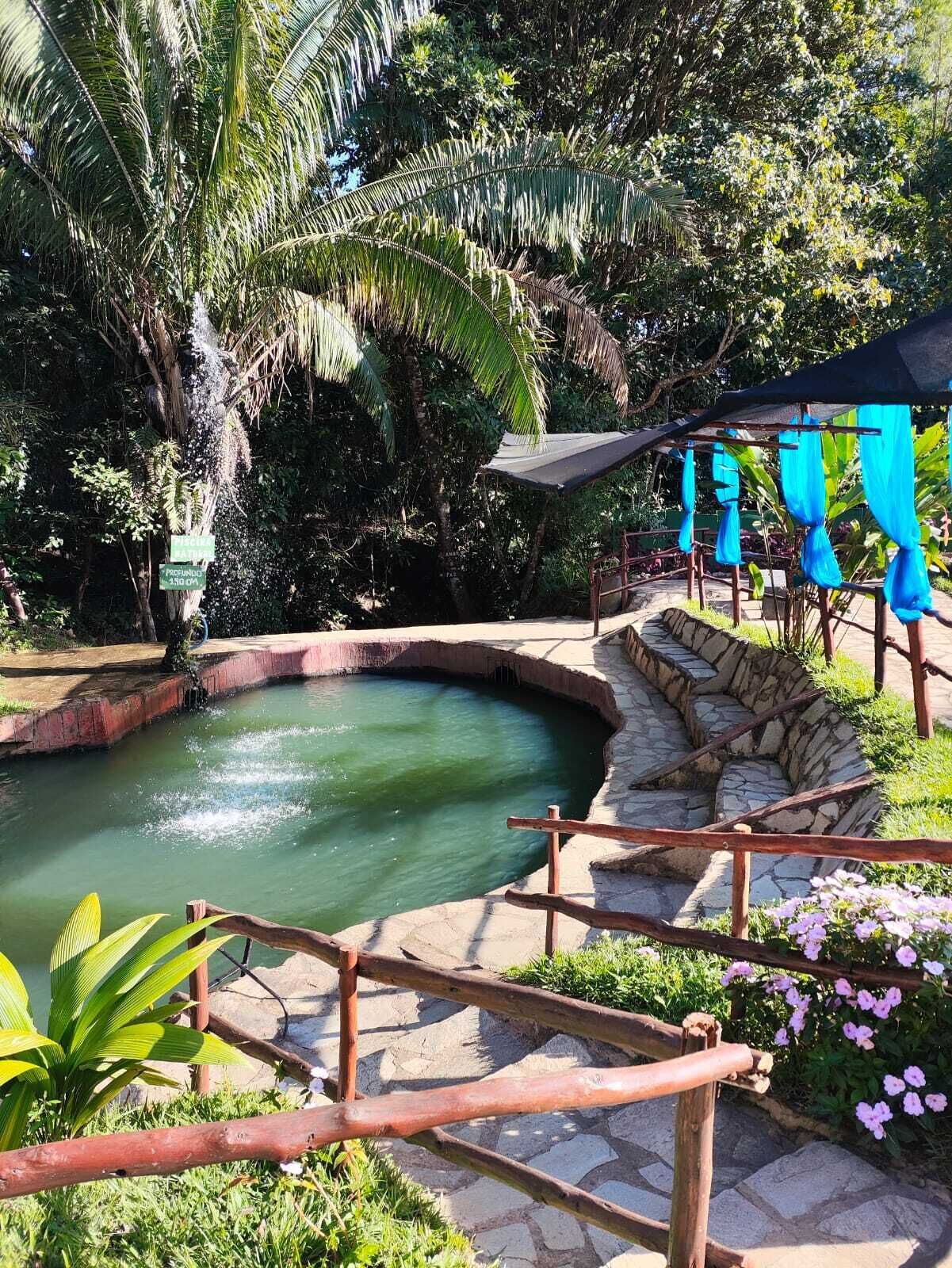 Uma piscina de pedra rodeada por vegetação exuberante, uma cerca de madeira e uma área de estar coberta por toldos azuis.
