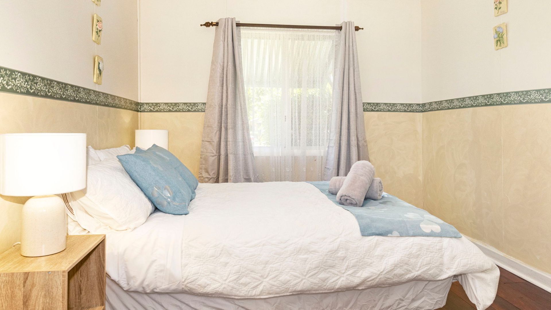 Een slaapkamer met een bed, twee kussens, een nachtkastje, een lamp en een raam met gordijnen.