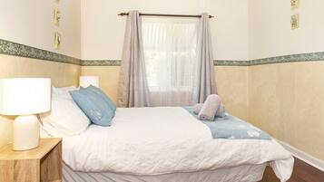 Een slaapkamer met een bed, twee kussens, een nachtkastje, een lamp en een raam met gordijnen.