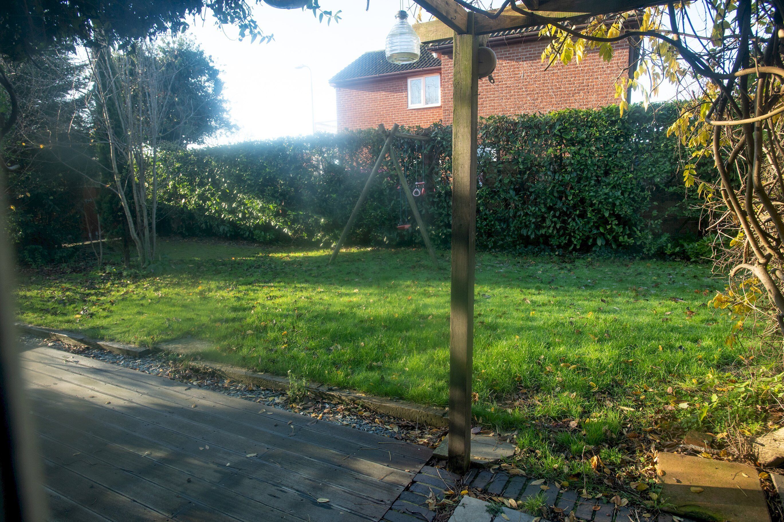 A residential garden with a wooden deck, a brick house, and a hedge.