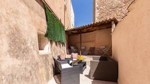 Un patio exterior estrecho con mobiliario de mimbre, una mesita con un florero y una pérgola encima.