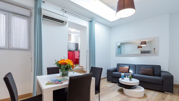 Una sala moderna con comedor, un sofá gris y una pequeña cocina visible a través de una puerta abierta.