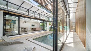Un área de piscina interior moderna con paredes de vidrio, una zona de estar con asientos y un comedor.