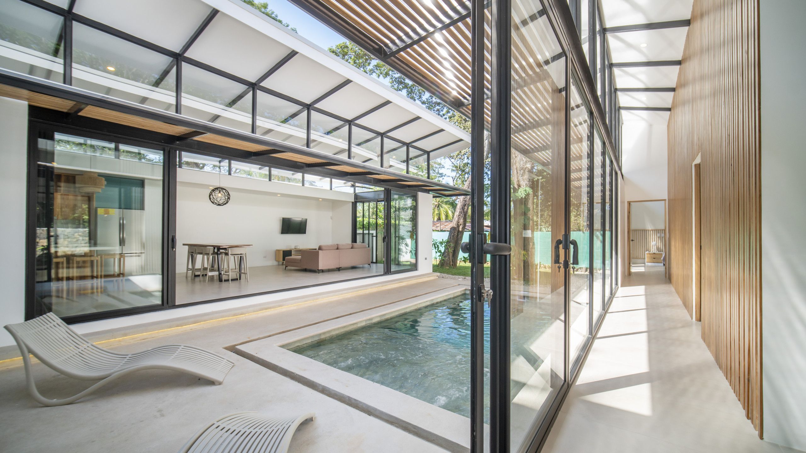 Un área de piscina interior moderna con paredes de vidrio, una zona de estar con asientos y un comedor.