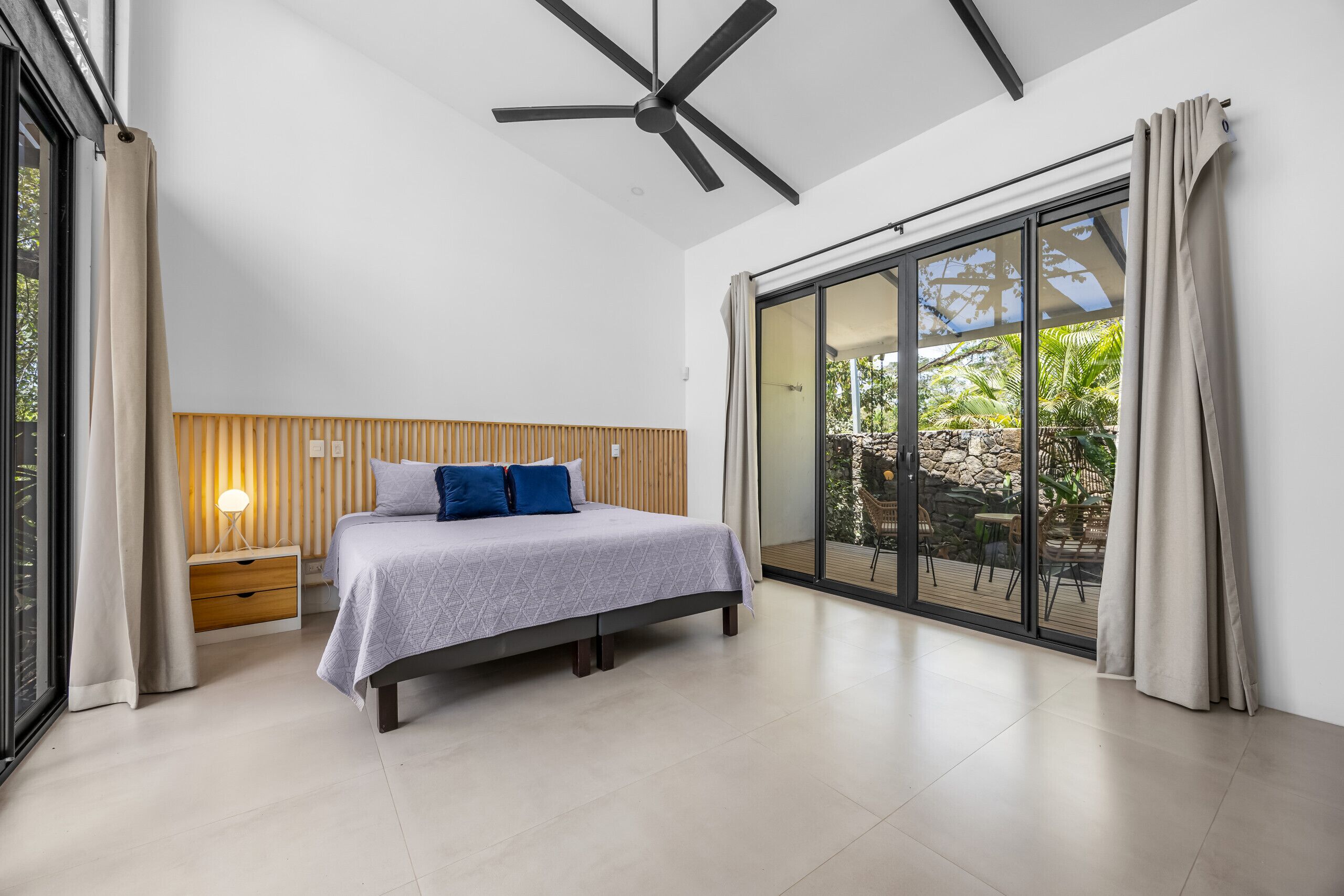 A modern bedroom with a bed, bedside table, lamp, ceiling fan, and large sliding glass door to an outdoor area.