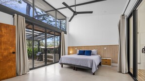 Un dormitorio moderno con una cama grande, un ventilador de techo y un ventanal corredero que da a un espacio exterior.
