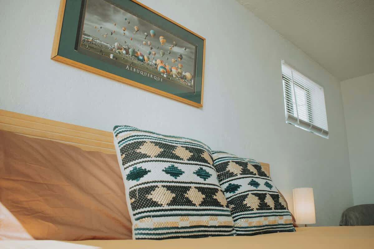 A hotel room with a bed, pillows, a framed picture on the wall, a window with blinds, and a lamp on the nightstand.