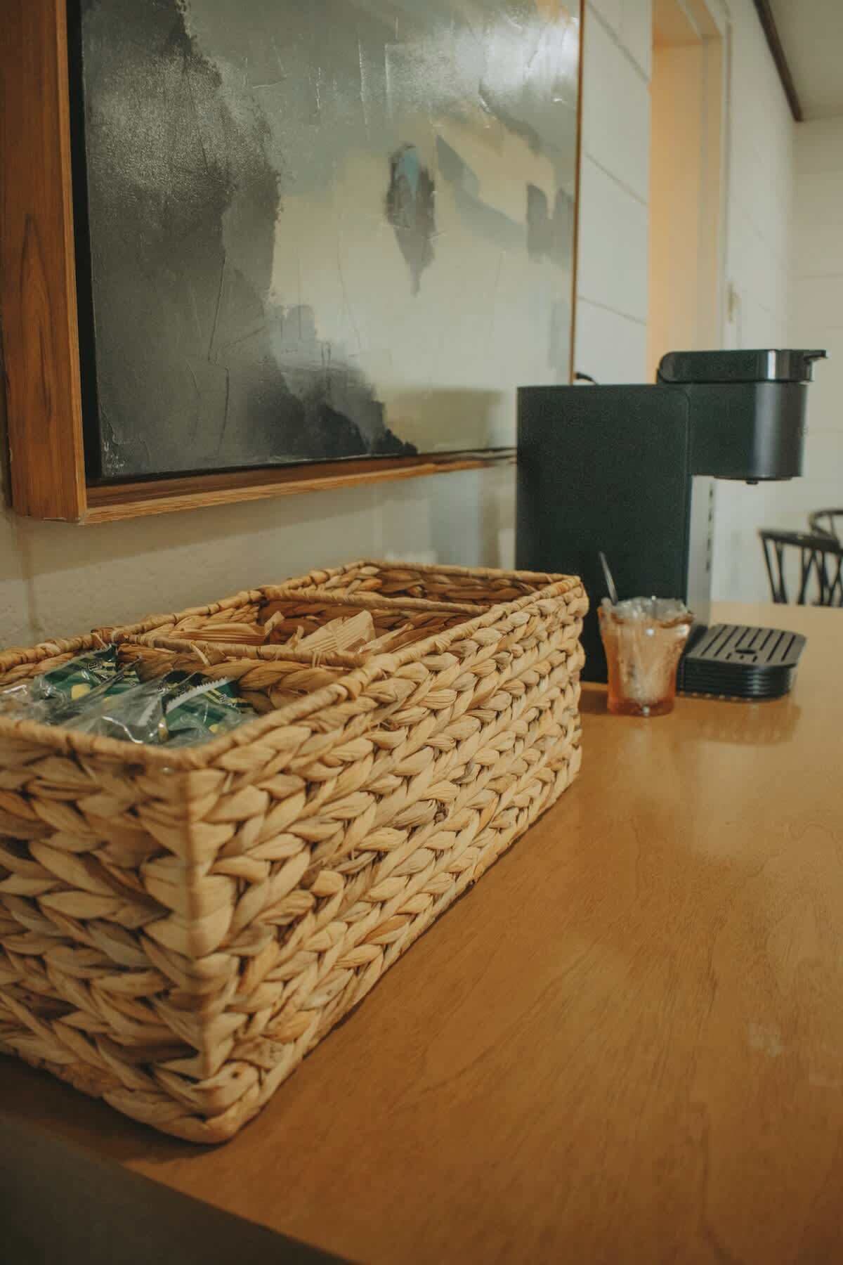 A woven basket on a wooden table with a glass of iced tea and a black object, possibly a kettle, in the background.