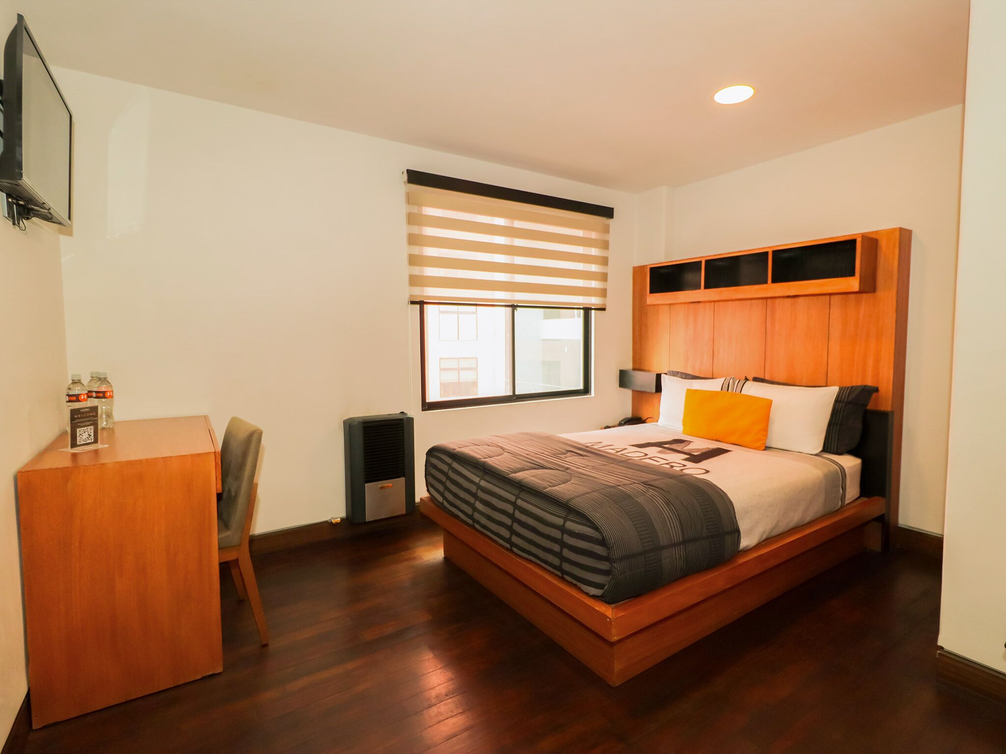 Habitación de hotel con cama de madera, un escritorio, una silla, ventana con persianas y televisor montado en la pared.
