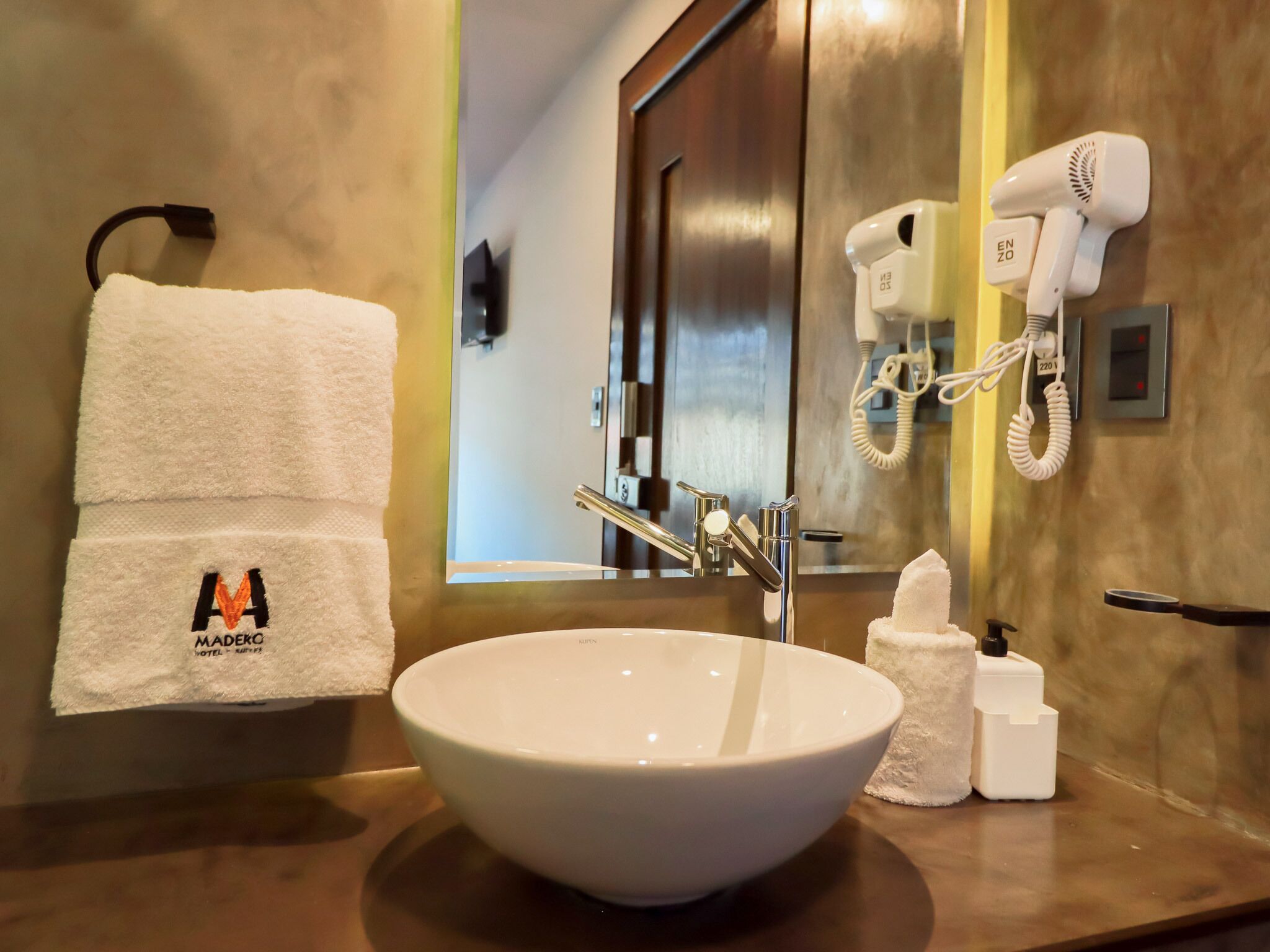 Ein Hotelbad mit einem weißen Waschbecken, einem Handtuch mit einem Logo, einem Fön und einem Spiegel.