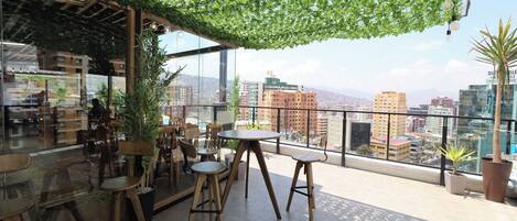 A rooftop terrace with wooden furniture, potted plants, and a view of the cityscape.