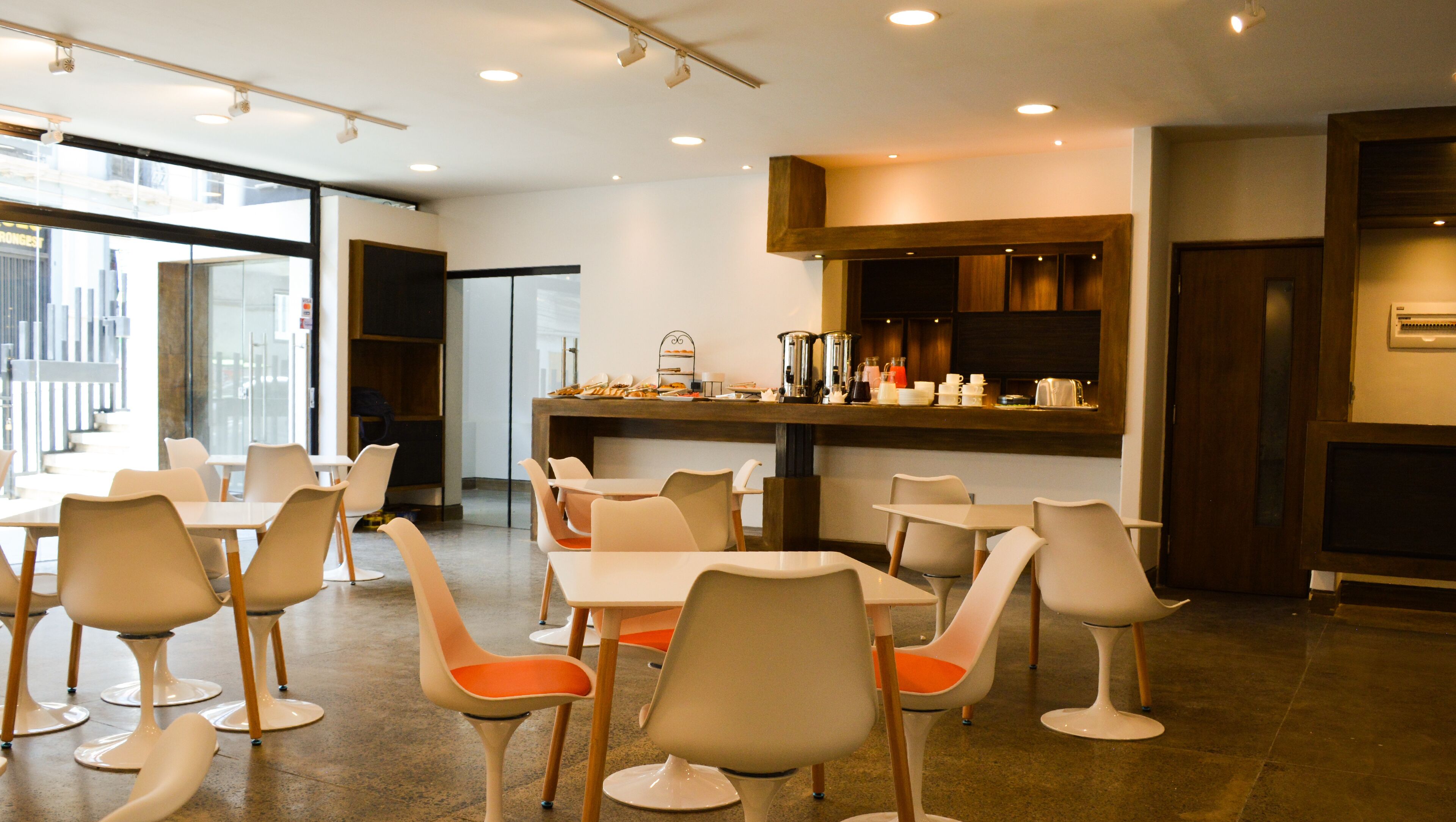 A modern dining area with white tables and chairs, a central bar area with a coffee station, and large windows providing natural light.