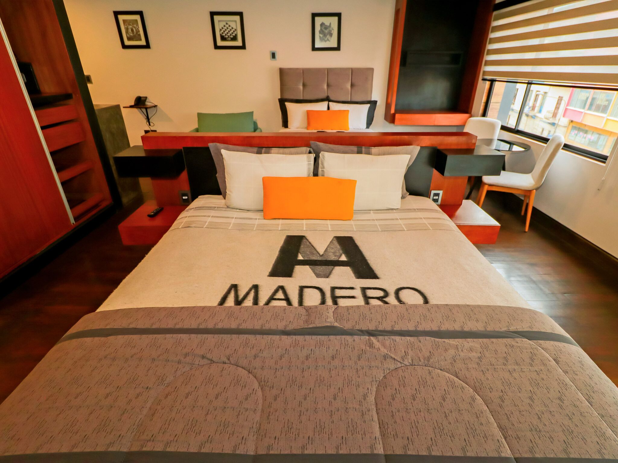 A hotel room with a bed featuring a large 'MADERO' logo, a wooden headboard, and a view of the city through the window.