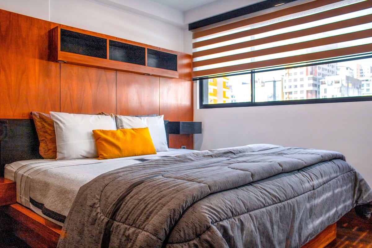 A modern bedroom with a large bed, wooden headboard, and a city view through a window with blinds.