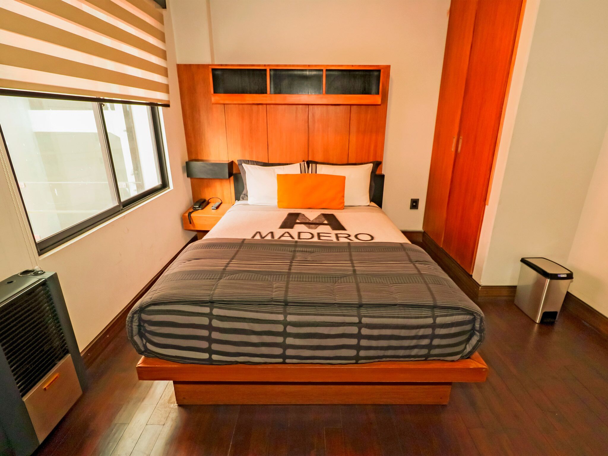 A modern bedroom with a wooden headboard, a bed with a striped mattress, a nightstand with a lamp, and a window with blinds.
