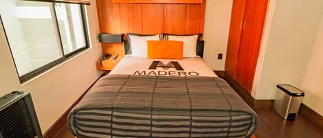 Een moderne slaapkamer met een houten hoofdbord, een bed met een gestreept matras, een nachtkastje met een lamp en een raam met zonwering.