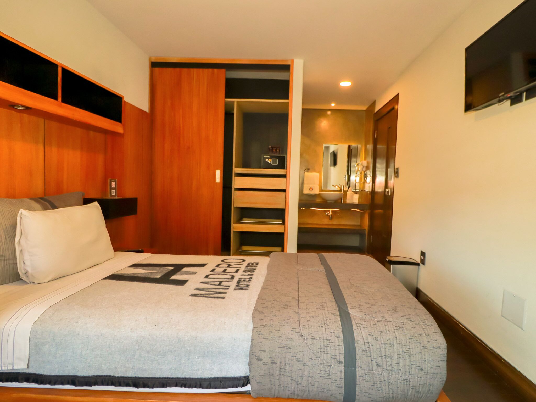 A hotel room with a bed, a wooden wardrobe, a bathroom with a sink and mirror, and a TV mounted on the wall.
